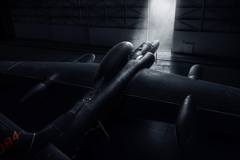 Dramatic low-angle view of a military aircraft fuselage illuminated by stark overhead lighting in a hangar.