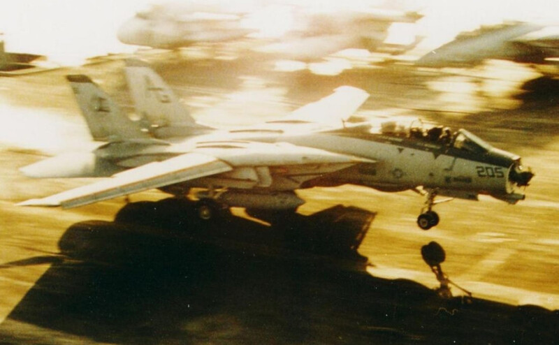 Military fighter jet in flight with weapons payload visible beneath fuselage.