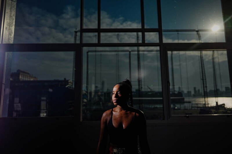 Woman in dark tank top positioned against large industrial windows with city skyline visible behind glass.