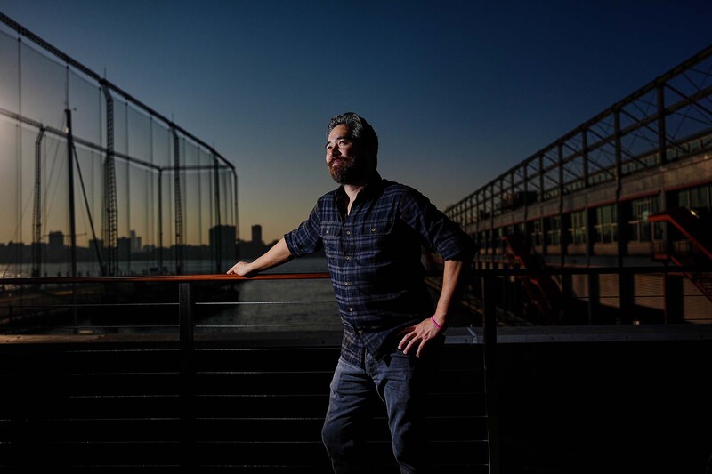 Man in plaid shirt leaning against railing on industrial rooftop at golden hour.