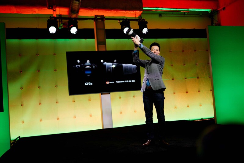 Presenter on stage demonstrating camera equipment with raised arm against colorful backdrop and display screen.