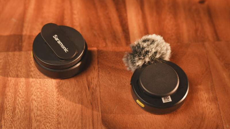 Two wireless microphone capsules with windscreen covers sit on a wooden surface.