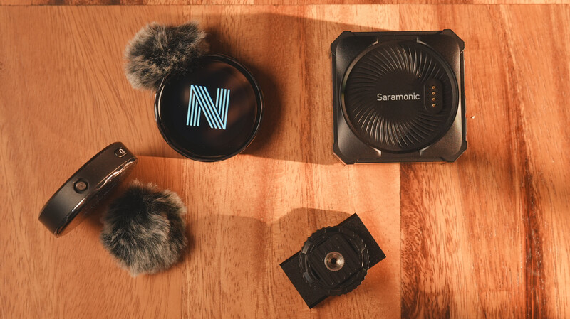 Four pieces of audio equipment arranged on a wooden surface: a wireless microphone system, a compact audio interface, and mounting accessories.