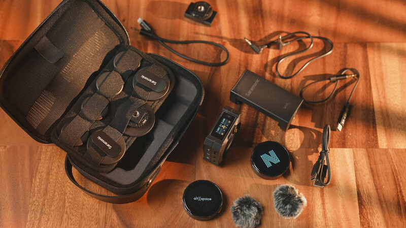 Professional audio and video gear laid out on a wooden surface, including microphones, windscreens, cables, and a carrying case.