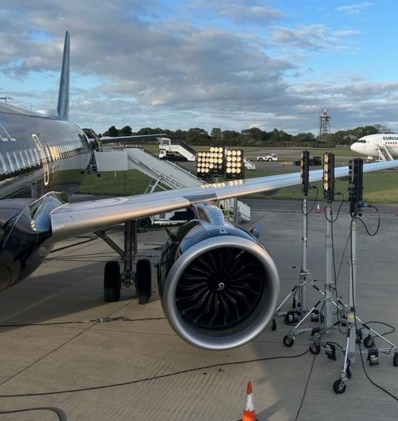 Commercial aircraft engine and fuselage on airport tarmac with ground support equipment and facilities visible.