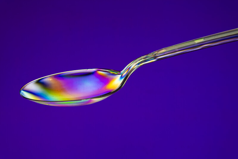 Clear spoon with iridescent oil film creating rainbow colors against deep purple background.