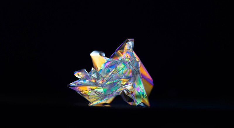 Translucent crystal or glass sculpture with iridescent rainbow refractions against a black background.