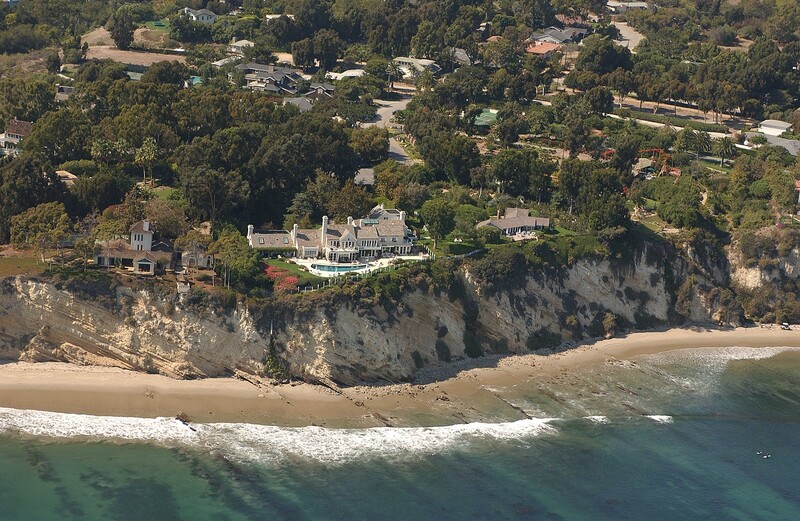 Aerial view of a large mansion on a coastal bluff overlooking a sandy beach and turquoise ocean.