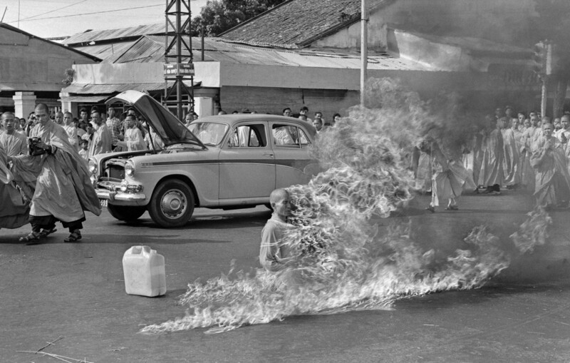 Person engulfed in flames during a public demonstration as onlookers and a parked car watch from a distance.