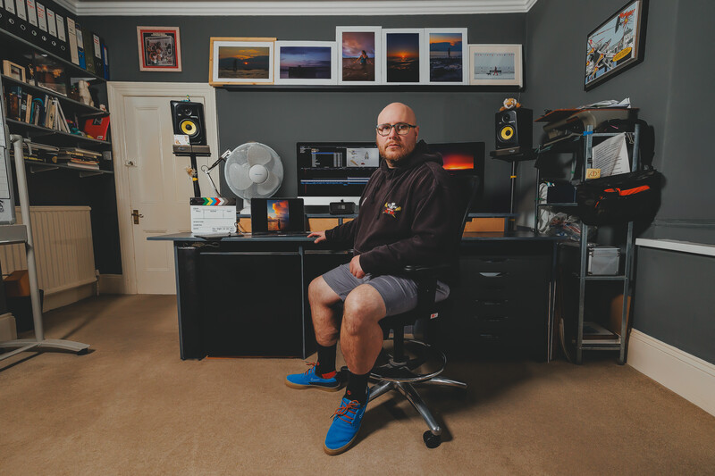 Man seated at desk in home studio setup with monitor, keyboard, and professional audio speakers.