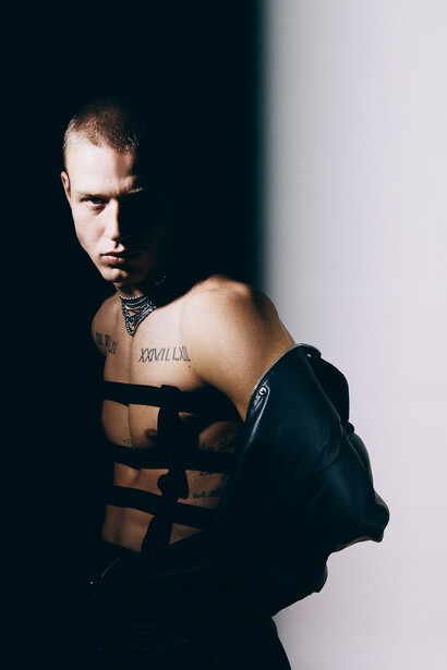 Shirtless man with geometric black tape strips across his torso, photographed against a split light and dark background.
