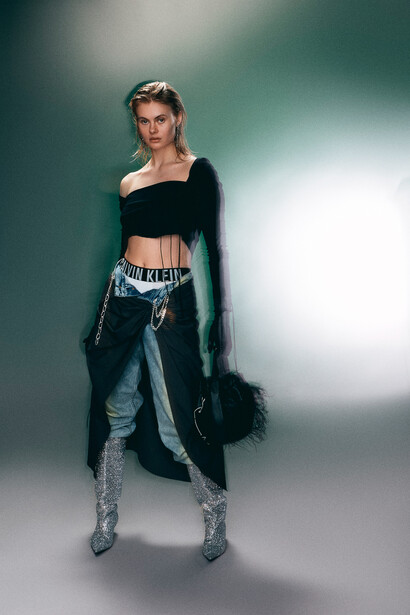 Woman in black off-shoulder crop top and patterned leggings poses against gradient teal and white backdrop.