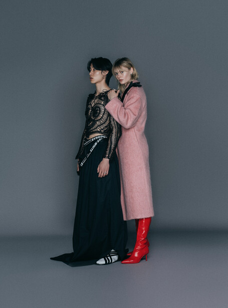 Two figures posed closely together against a neutral gray backdrop, one wearing a black sequined outfit and the other in a pink coat with red boots.