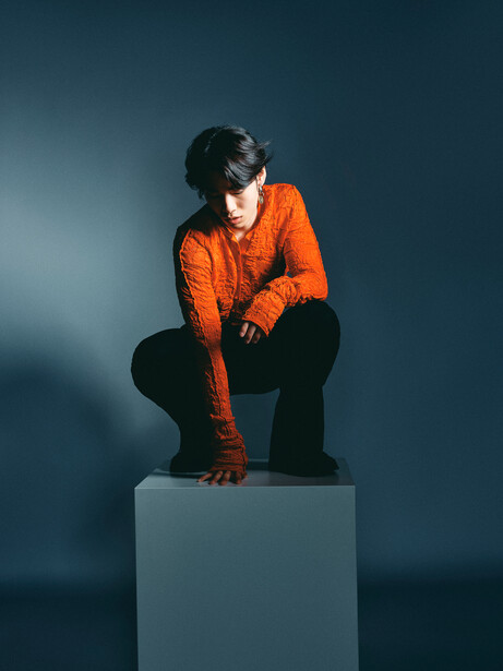 Person in bright orange jacket crouching on gray cube against blue background.