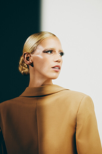 Woman with blonde hair styled in a low bun, wearing a mustard-colored long-sleeved top, looking upward against a split dark and light background.