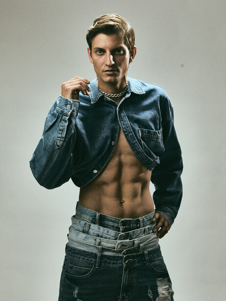 Shirtless male model wearing open denim jacket and distressed jeans against neutral background.
