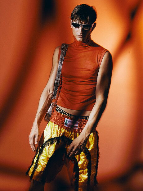 Young man wearing red sleeveless shirt and yellow patterned pants against warm orange background.