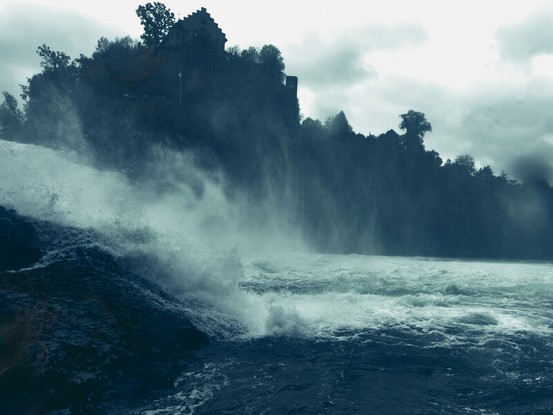 Turbulent river with massive waves crashing against rocky banks beneath a misty, forested cliff.