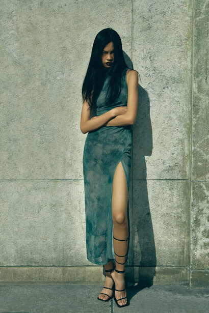 Woman with long dark hair wearing a teal velvet dress with high slit against textured concrete wall.