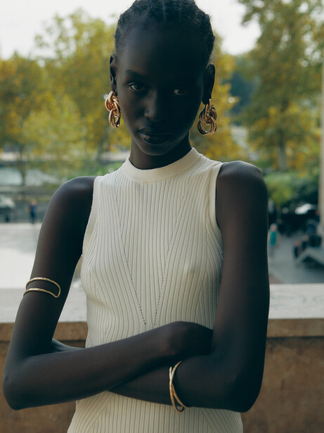 Portrait of a woman with arms crossed, wearing a white sleeveless ribbed top and gold jewelry, photographed outdoors.