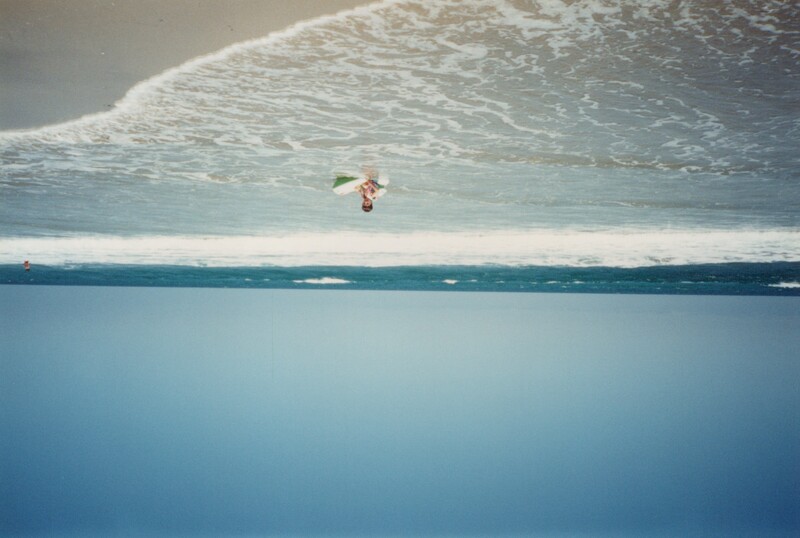 Surfer in shallow water holding a white surfboard, standing in gentle waves along a sandy beach.