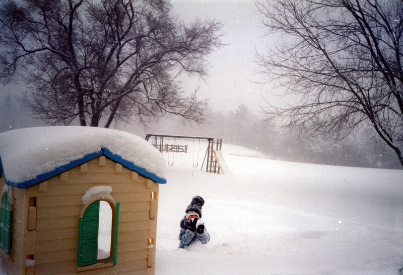 Child sledding in deep snow near wooden playground structures on an overcast winter day.