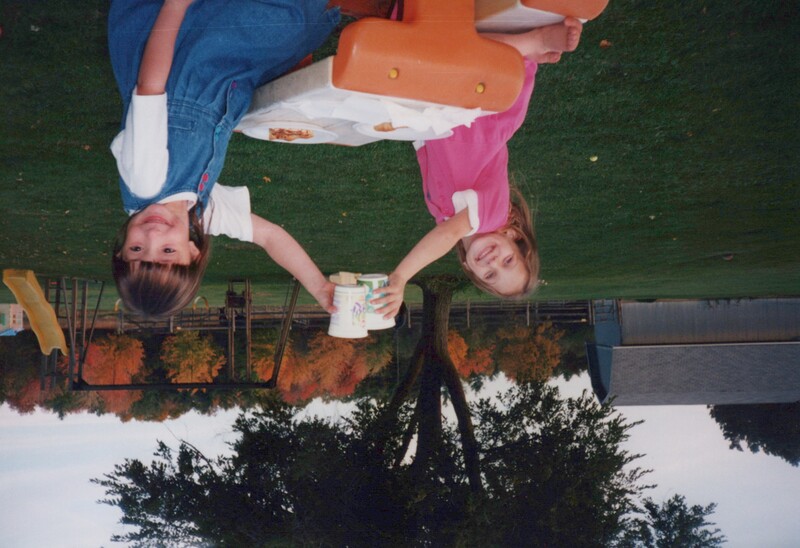 Two young girls sitting at a picnic table outdoors, holding up a plate of food between them.