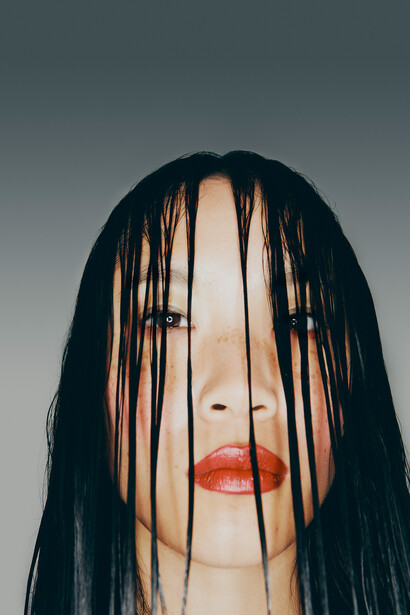 Portrait of a person with long black hair partially covering their face, wearing bold red lipstick against a neutral gray background.