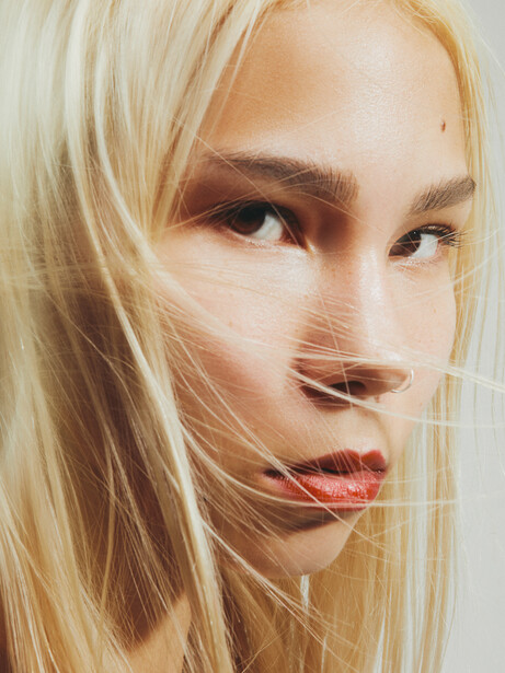 Portrait of a blonde woman with neutral expression and subtle eye makeup against a light background.