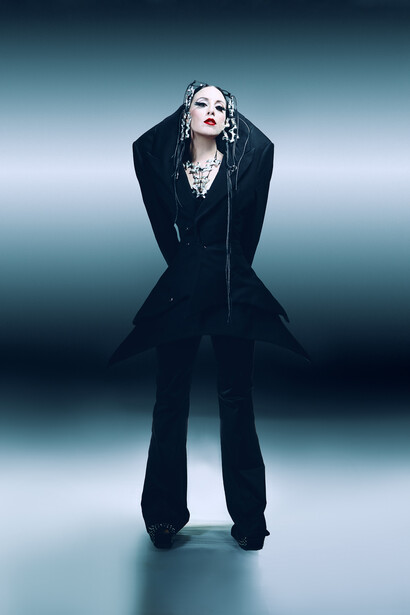Person in black tailored suit standing against gradient blue-gray backdrop with dramatic lighting.