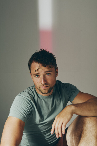 Man in gray t-shirt seated against neutral wall with warm window light casting soft shadows.