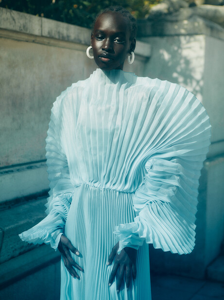 Person in light blue pleated dress with matching headwrap against concrete wall in natural daylight.