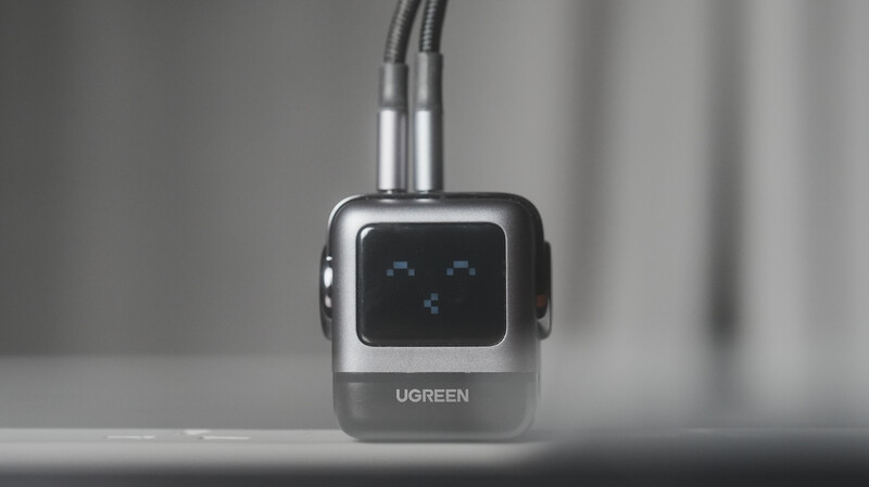Ugreen USB hub with square black display screen and curved cable connection against neutral gray background.