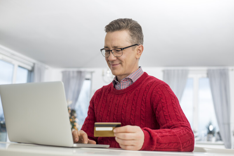 Man in red cable-knit sweater holding credit card while looking at laptop screen.
