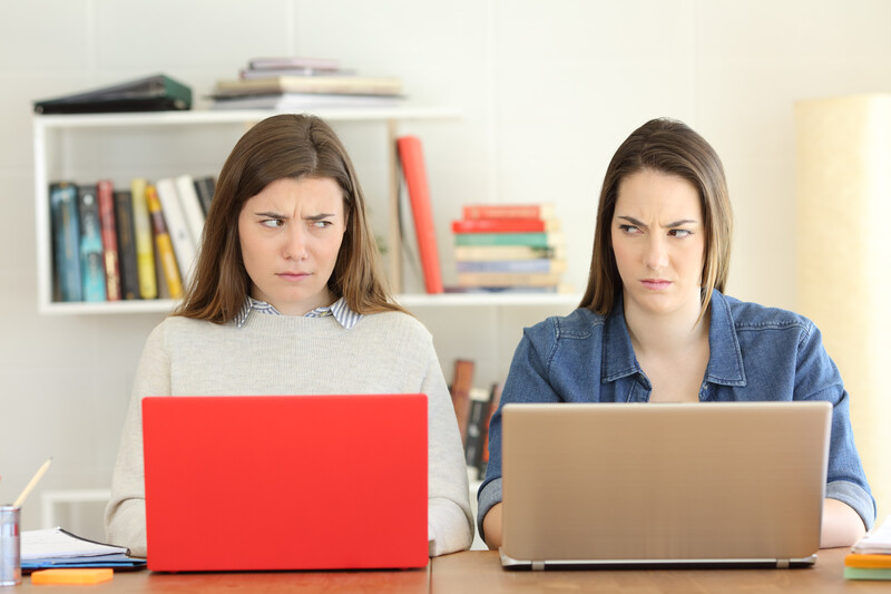 Two women sitting at a desk with laptops, both with serious expressions looking directly at camera.