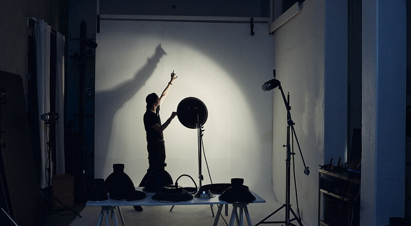 Silhouetted figure adjusting a large reflector on a studio set with dramatic backlighting.