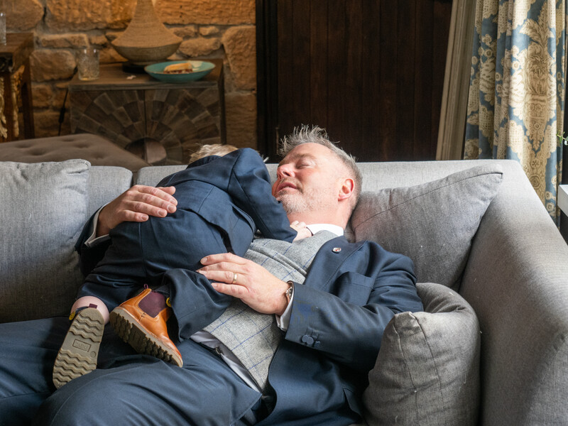 Man in navy blazer reclining on gray sofa, holding and looking down at sleeping child in dark clothing.