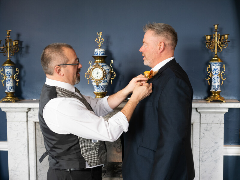 Man adjusting tie on another man's formal attire against blue wall backdrop.