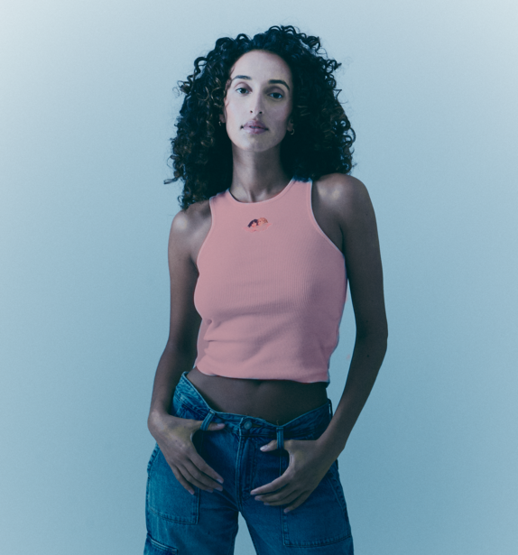 Woman with curly dark hair wearing pink sleeveless top and blue jeans against light blue background.