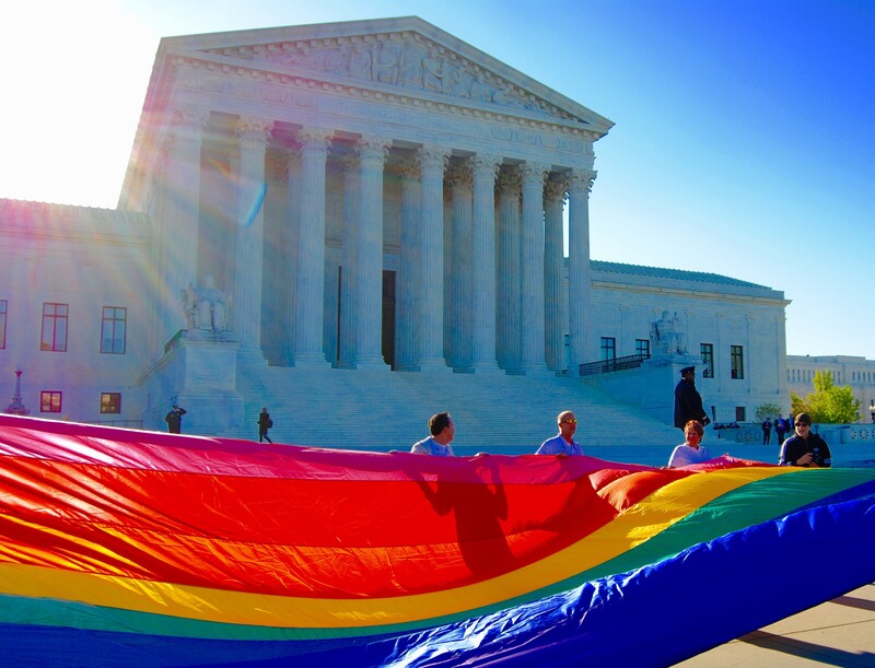 Large rainbow pride flag held by people in front of a classical government building with white columns.