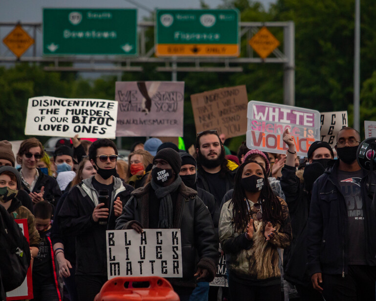 Large crowd of protesters holding signs on a highway overpass beneath green directional road signs.