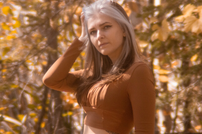 Woman with long gray hair posing outdoors among golden autumn foliage.