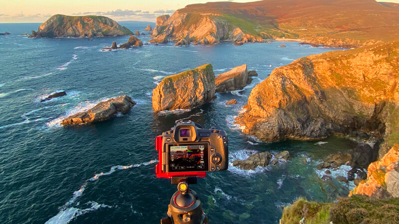Red mirrorless camera mounted on tripod overlooking rocky coastal cliffs and islands at golden hour.