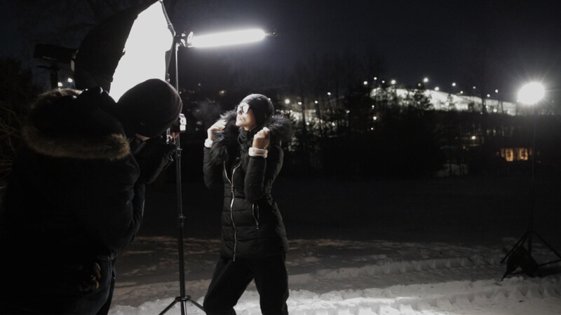 Portrait subject posed under professional lighting setup during nighttime outdoor shoot with city lights in background.