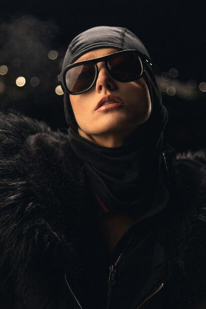 Person wearing dark sunglasses and helmet against blurred nighttime background with bokeh lights.