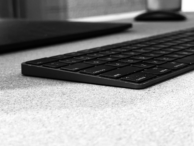 Black wireless keyboard on light textured surface with blurred desk items in background.