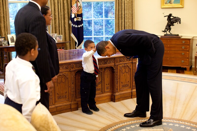 An adult bends down to interact with a young child in an ornate formal room with classical wood paneling.