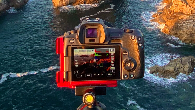 DSLR camera mounted on tripod displaying coastal landscape with rocky cliffs and turquoise water on LCD screen.