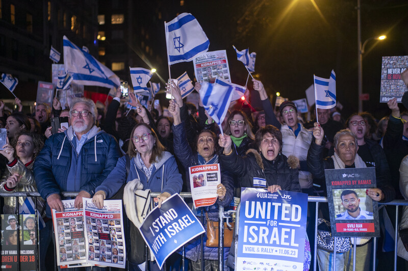 Nighttime crowd holding Israeli flags and signs at an outdoor rally or demonstration.