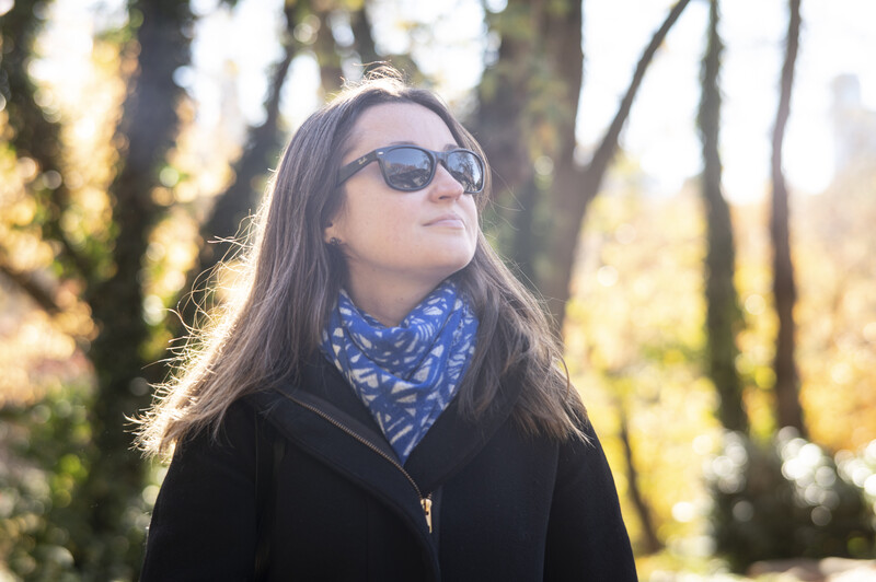 Woman wearing sunglasses and dark jacket outdoors among trees in golden sunlight.